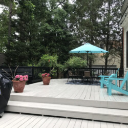 A light-colored wooden deck with a grill, potted flowers, a patio table with an umbrella, and two Adirondack chairs.