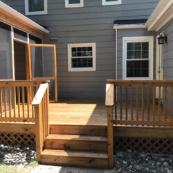 A wooden deck with stairs and railings attached to the back of a grey house.
