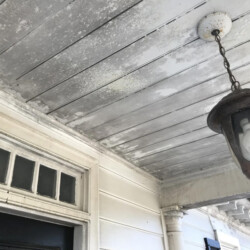 A metal porch light hanging from a weathered, paint-peeling wooden ceiling above a front door.
