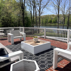 A brick patio with white outdoor furniture and a central coffee table, overlooking a lake surrounded by trees.