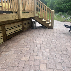 A red brick patio beneath a wooden deck with stairs and a lounge chair.