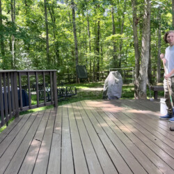 A young man stands on a wooden deck holding a paint roller, with trees in the background.