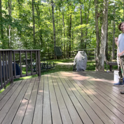 A young man stands on a wooden deck holding a paint roller, with a forest in the background.
