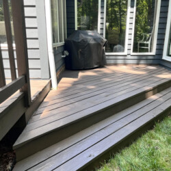 A grey wooden deck with stairs leading to a covered grill outside a house.