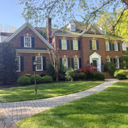 A two-story brick house with a paved walkway, front lawn, and trees.