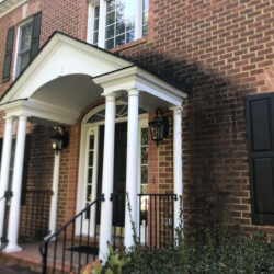 A brick house exterior with a white portico, black shutters, and dark staining on the masonry near the entrance.