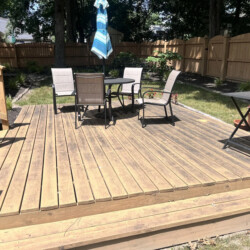 Wooden deck with a table and four chairs, a closed patio umbrella, and a side table, enclosed by a wooden fence.