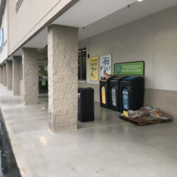 A row of recycling and trash bins outside a store entrance, next to a wooden pallet holding bags of mulch.