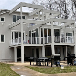 Three-story modern home with light gray exterior, balconies, and a ground-level patio with dining furniture and grills.