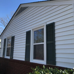 Exterior view of a house with white horizontal siding, two windows with dark shutters, and a brick base.