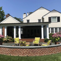 Patio with four chairs and a curved brick wall in front of a house with a covered porch.
