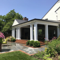 A covered brick patio with white columns attached to a brick house, surrounded by a landscaped garden and lawn.