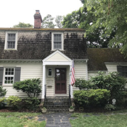 Two-story Dutch Colonial house with white siding, dark shutters, and an American flag by the front door.