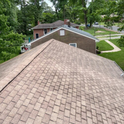 High-angle view of a residential shingle roof with a brick house and suburban yard in the background.