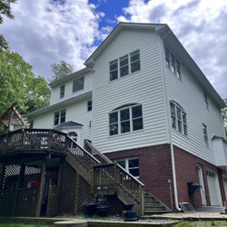 Three-story house with white siding and a brick lower level, featuring a wooden deck and staircase.